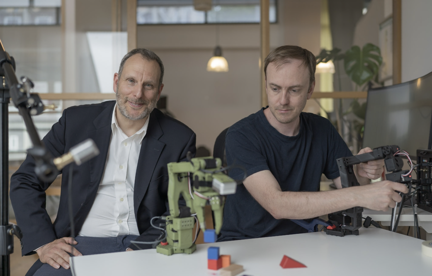 Two men working with a robot arm at a desk. Computers in the background.