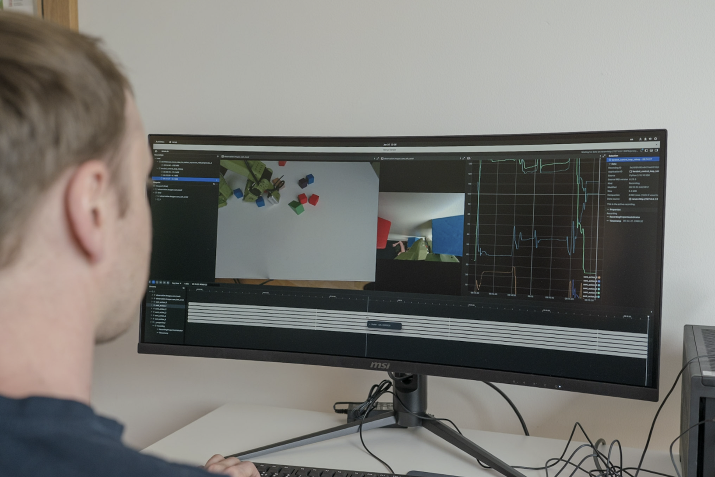 A man in front of a screen showing camera-on-hand robot view and top camera view, and graph of robot joint angle values.