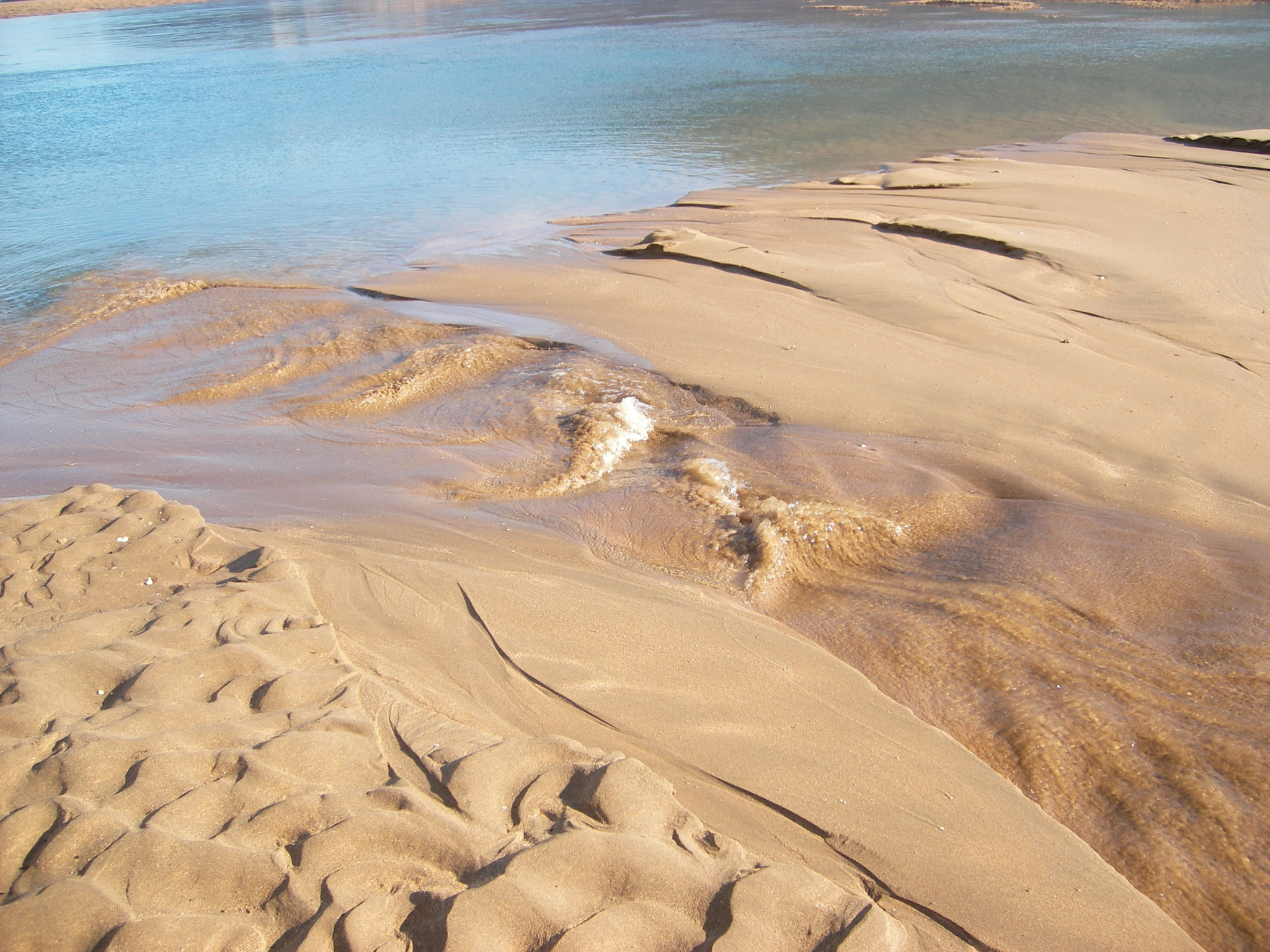 A high-angle photograph showing water runoff cutting through a sandy beach slope, creating small rippled channels as it empties into a larger body of clear blue water.