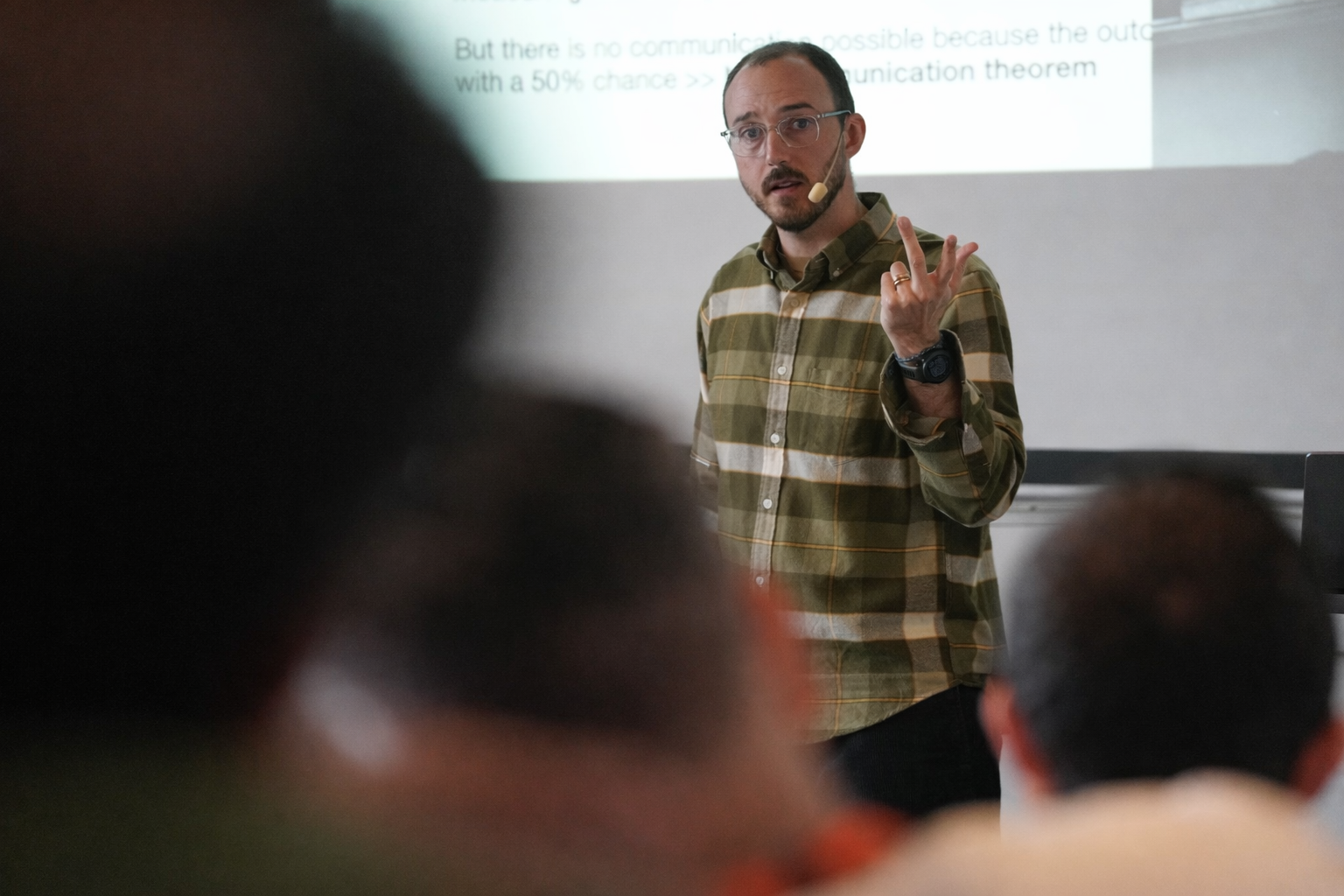 A speaker wearing a headset microphone gives a presentation in front of an audience, gesturing while standing beside a projected slide.
