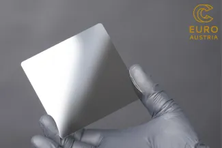 A human hand in a sterile glove, holding a 10x10 cm ceramic tablet between thumb and trigger finger. All held in grey colour.
